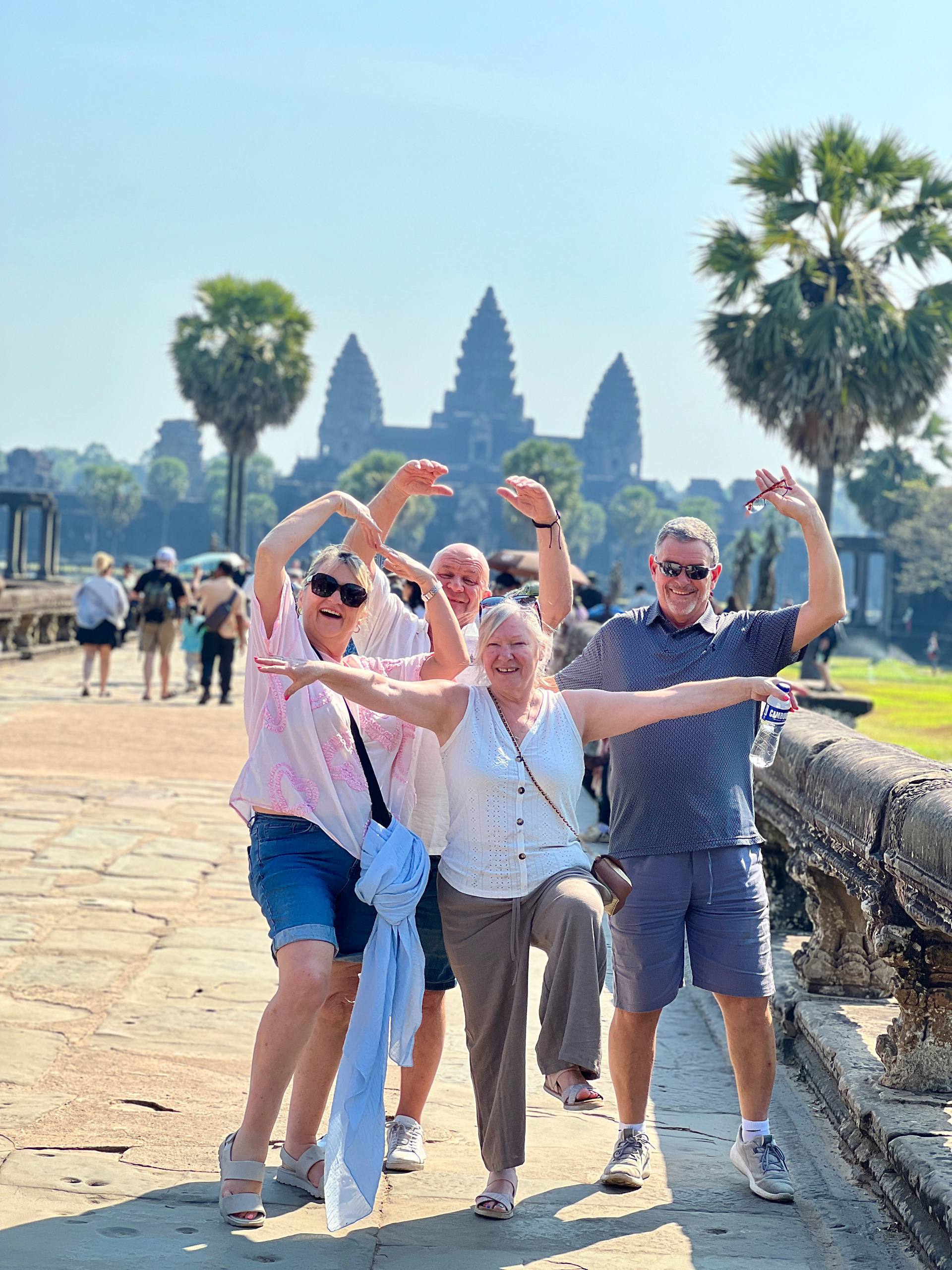 Four smiling tourists posing playfully on the stone causeway in front of Angkor Wat temple.